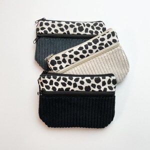 Könnte beinhalten: Drei kleine Reißverschlusstaschen in einem Stapel. Die Taschen haben eine Kordstruktur in Schwarz, Grau und Beige. Jede Tasche hat einen oberen Bereich mit einem schwarz-weißen Fleckenmuster. Die Reißverschlüsse sind schwarz.