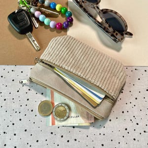 May include: A beige corduroy zippered pouch with a light tan base, open to reveal cards and currency. A set of keys, a beaded bracelet, and tortoiseshell sunglasses are also visible.