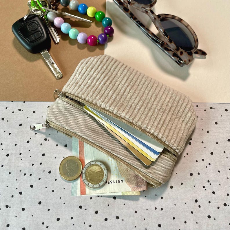 May include: A beige corduroy zippered pouch with a light tan base, open to reveal cards and currency. A set of keys, a beaded bracelet, and tortoiseshell sunglasses are also visible.