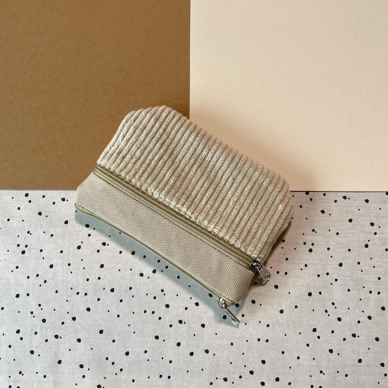 May include: A beige corduroy zippered pouch with a rectangular shape. The top section is made of ribbed corduroy, while the bottom is a smooth, matching fabric. The zipper is silver-toned. The pouch is on a speckled surface.