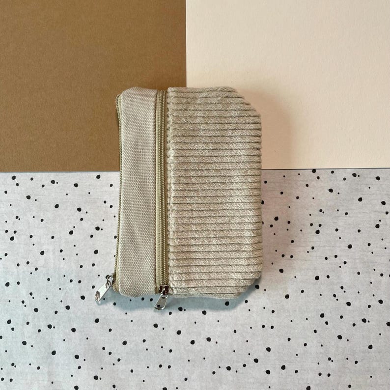 May include: A beige corduroy zippered pouch with a rectangular shape. The pouch has a lighter beige canvas-like section on the left side. The background is a mix of brown, beige, and a speckled pattern.