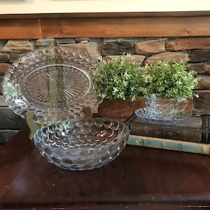 Puede incluir: Un juego de platos de vidrio transparente con textura de burbujas. El juego incluye una fuente ovalada grande para servir, un tazón redondo más pequeño y una taza de té y un platillo.