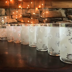 Puede incluir: Conjunto de ocho vasos de vidrio esmerilado con un diseño de copo de nieve. Los vasos están dispuestos en una fila sobre una superficie de madera oscura.