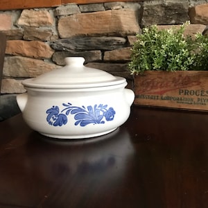 Puede incluir: Una fuente de cerámica blanca con tapa, con diseños florales azules. La fuente tiene dos asas y está sobre una superficie de madera oscura. Una caja de madera con vegetación está en el fondo.