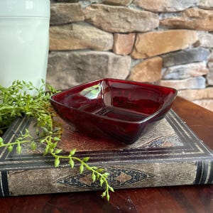 May include: A square, deep red glass bowl with smooth, rounded corners and a polished finish. The bowl features a translucent, dark ruby hue that catches the light, showcasing a simple, open design with slightly flared sides and a flat base.