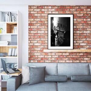 Op de afbeelding: Een zwart-witfoto van een stadssc&egrave;ne met mensen die op de voorgrond lopen. De foto is ingelijst in een zwart frame en hangt aan een bakstenen muur. De muur is geschilderd in tinten rood en oranje. Een grijze bank met kussens staat op de voorgrond.