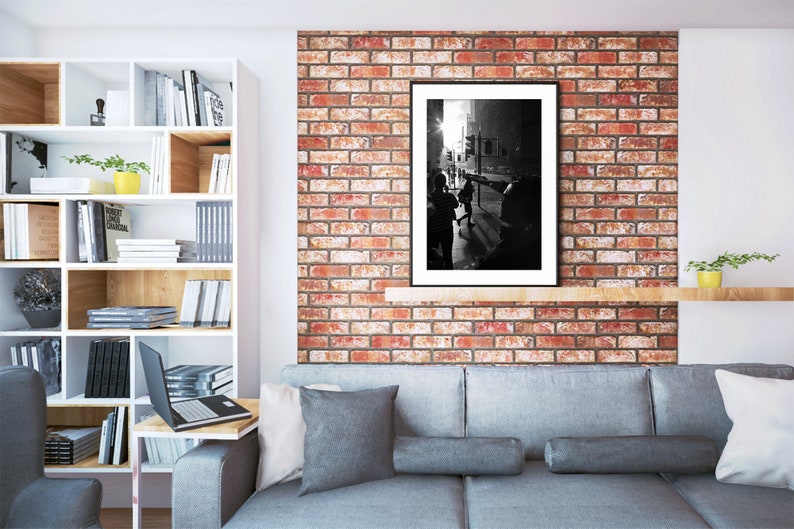 Op de afbeelding: Een zwart-witfoto van een stadssc&egrave;ne met mensen die op de voorgrond lopen. De foto is ingelijst in een zwart frame en hangt aan een bakstenen muur. De muur is geschilderd in tinten rood en oranje. Een grijze bank met kussens staat op de voorgrond.