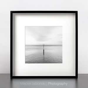 Puede incluir: Una fotografía en blanco y negro de un solo poste en el agua. El poste es una simple estructura de metal con una parte superior cuadrada. El agua está tranquila y el cielo está nublado. La foto está enmarcada en un marco negro.