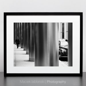 May include: A black and white photograph of a person walking down a city street lined with tall, shiny columns. A black London taxi cab is parked on the right side of the image, partially obscured by a column.