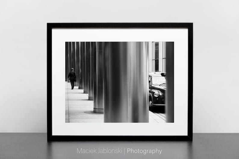 May include: A black and white photograph of a person walking down a city street lined with tall, shiny columns. A black London taxi cab is parked on the right side of the image, partially obscured by a column.