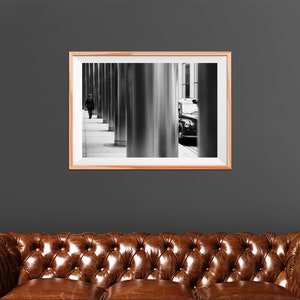 May include: Black and white photograph of a person walking down a city street with a black car parked in the background. The photo is framed in a light wood frame.