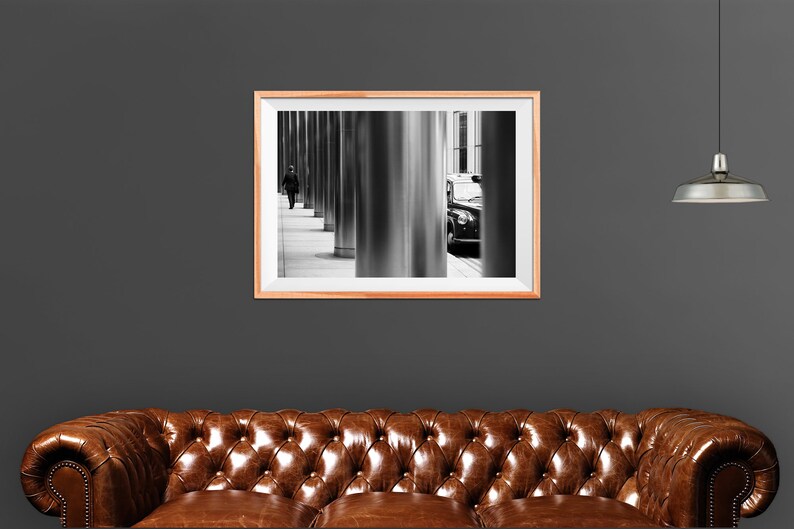 May include: Black and white photograph of a person walking down a city street with a black car parked in the background. The photo is framed in a light wood frame.
