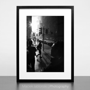 Op de afbeelding: Zwart-witfoto van mensen die over een straat in de stad lopen. De zon schijnt fel en de straat is nat van de regen. De afbeelding is ingelijst in een zwart frame.