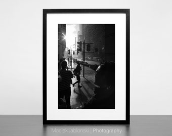 May include: Black and white photograph of people walking down a city street. The sun is shining brightly and the street is wet from rain. The image is framed in a black frame.