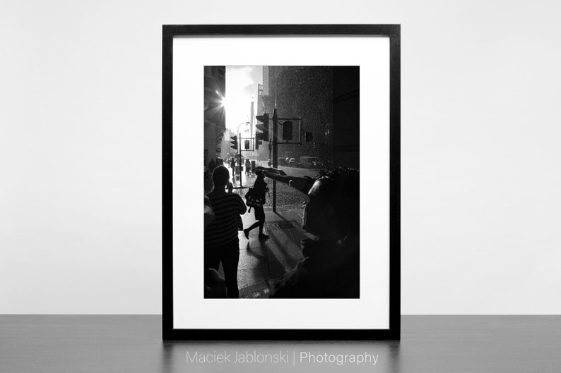 Op de afbeelding: Zwart-witfoto van mensen die over een straat in de stad lopen. De zon schijnt fel en de straat is nat van de regen. De afbeelding is ingelijst in een zwart frame.
