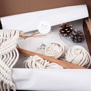 K&ouml;nnte beinhalten: Ein Makramee-Wandbehang-Set mit wei&szlig;em Baumwollseil, einem Holzstab und Anleitungen. Das Set ist in einem braunen Karton mit wei&szlig;em Seidenpapier verpackt.