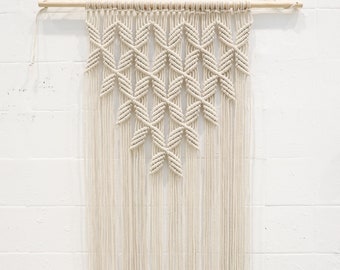 Peut inclure: Une suspension murale en macramé blanche avec un motif géométrique. La suspension est faite de cordon de coton et a un bâtonnet en bois en haut.