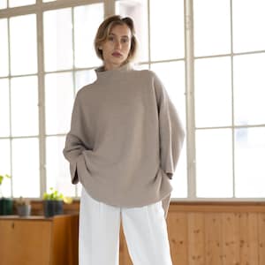 Könnte beinhalten: Eine Frau trägt einen hellbraunen Rollkragenpullover mit lockerer Passform und eine weiße, weite Hose. Sie steht in einem Raum mit großen Fenstern und Holzwänden.