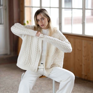 Può includere: Un cardigan bianco in maglia a treccia con chiusura a bottone. Il cardigan è indossato sopra una maglietta bianca e pantaloni bianchi. La persona è seduta su una sedia bianca.