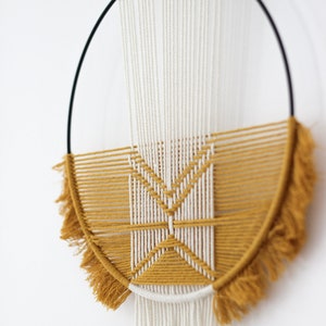 Pode incluir: Uma tapeçaria de macramé de parede com uma moldura circular de madeira e um padrão geométrico em fio amarelo mostarda e branco. A tapeçaria tem franjas longas brancas.