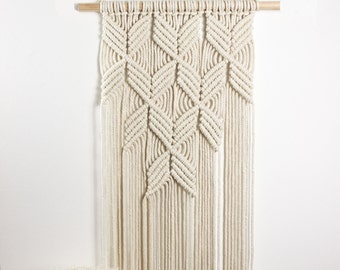 May include: A white macrame wall hanging with a geometric pattern. The hanging is made of cotton cord and has a wooden dowel at the top.