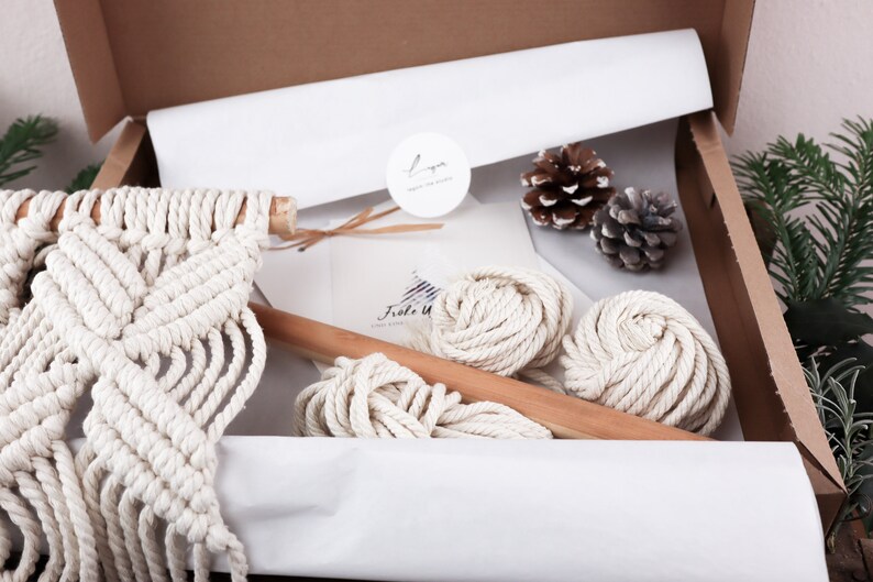 K&ouml;nnte beinhalten: Ein Makramee-Wandbehang-Set mit wei&szlig;em Baumwollseil, einem Holzstab und Anleitungen. Das Set ist in einem braunen Karton mit wei&szlig;em Seidenpapier verpackt.