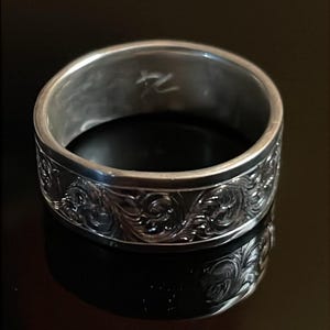 May include: A silver-toned metal ring featuring an intricate, embossed scrollwork pattern around the exterior band. The interior of the ring displays a small, etched symbol that resembles the characters 74 or a stylized mark.