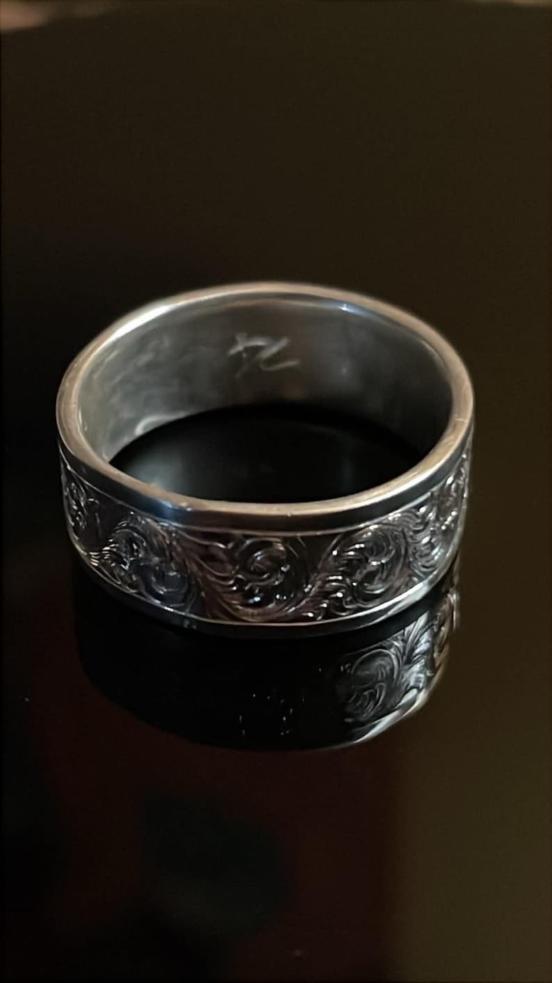 May include: A silver-toned metal ring featuring an intricate, embossed scrollwork pattern around the exterior band. The interior of the ring displays a small, etched symbol that resembles the characters 74 or a stylized mark.