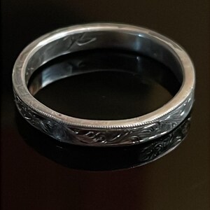 May include: A silver-toned metal ring featuring a continuous engraved floral scroll pattern along the outer band. The ring has a polished finish with a thin beaded border detail along the top and bottom edges and an inscription inside the band.