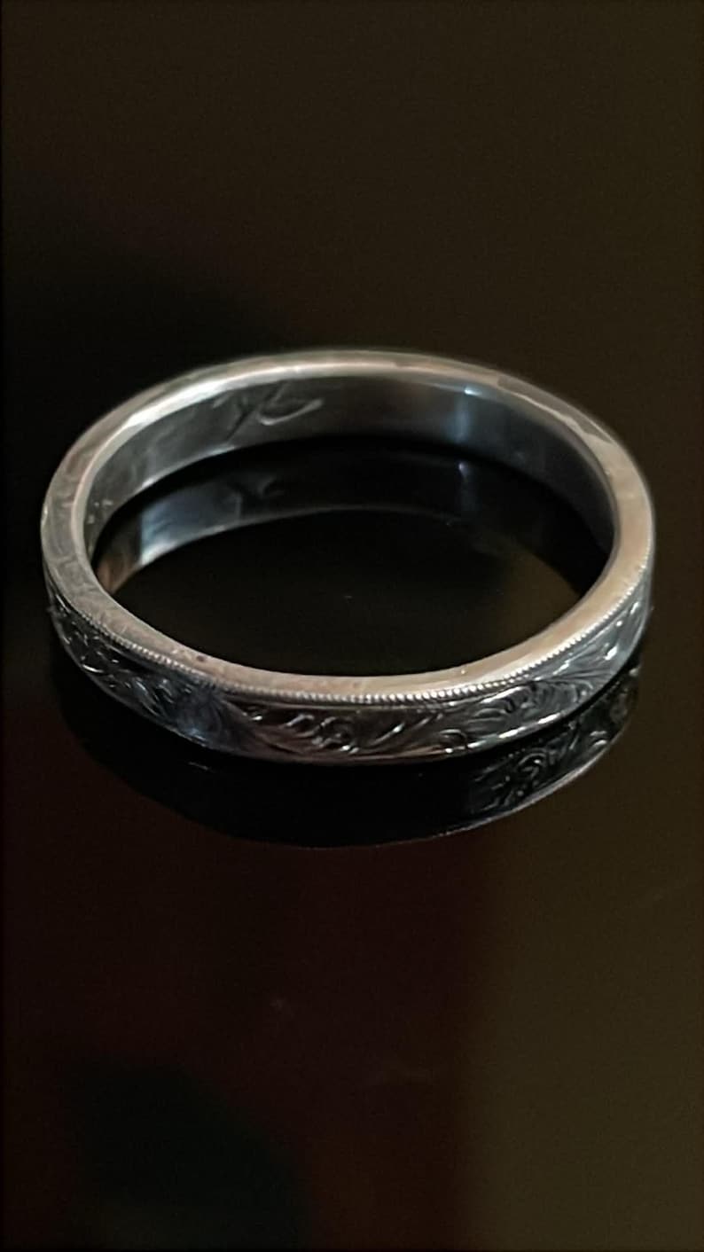 May include: A silver-toned metal ring featuring a continuous engraved floral scroll pattern along the outer band. The ring has a polished finish with a thin beaded border detail along the top and bottom edges and an inscription inside the band.