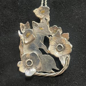 May include: Silver fox pendant necklace with gold detailing. The pendant depicts a fox amidst flowers. The necklace has a fine silver chain. The fox's eyes and flower centres are gold. The pendant is approximately 4 cm in length.