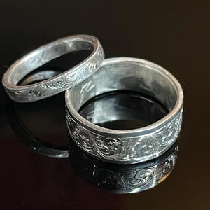 May include: Two silver-toned rings featuring intricate, engraved scrollwork patterns across their outer surfaces. The rings vary in width, with one being significantly thicker than the other, both displaying a polished metallic finish and detailed carvings.