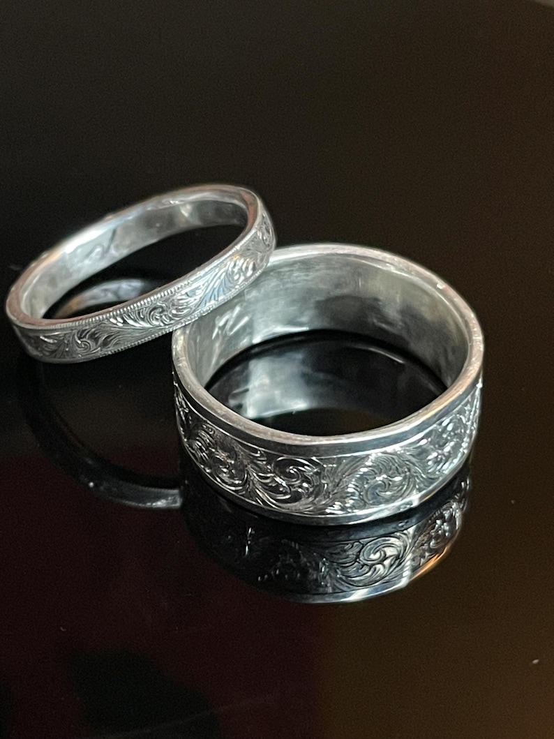 May include: Two silver-toned rings featuring intricate, engraved scrollwork patterns across their outer surfaces. The rings vary in width, with one being significantly thicker than the other, both displaying a polished metallic finish and detailed carvings.
