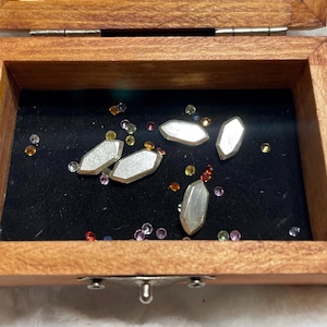 May include: A wooden box with a hinged lid, containing several silver-coloured, geometric-shaped objects and scattered colourful gemstones. The interior of the box is lined with black fabric, creating a contrasting backdrop for the items.