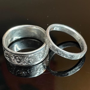 May include: Two silver-toned rings featuring intricate, engraved scrollwork patterns across their outer bands. The wider ring is positioned to the left, while the narrower ring sits to the right, both displaying a polished, reflective metallic finish.