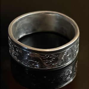 May include: A silver-toned metal ring featuring a wide band with a continuous embossed scrollwork pattern. The exterior surface of the band displays intricate, raised swirling designs that wrap around the entire circumference of the ring.