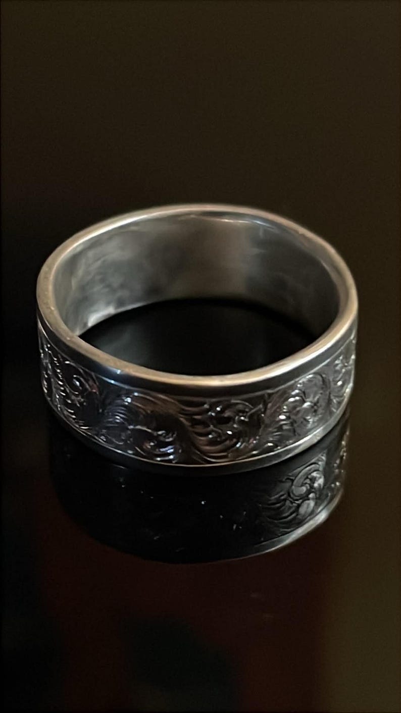 May include: A silver-toned metal ring featuring a wide band with a continuous embossed scrollwork pattern. The exterior surface of the band displays intricate, raised swirling designs that wrap around the entire circumference of the ring.