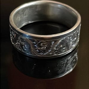 May include: A silver-toned metal ring featuring a continuous band with an intricate, embossed scrollwork pattern wrapping around the exterior surface. The ring has a smooth, polished interior and a reflective finish that catches the light.
