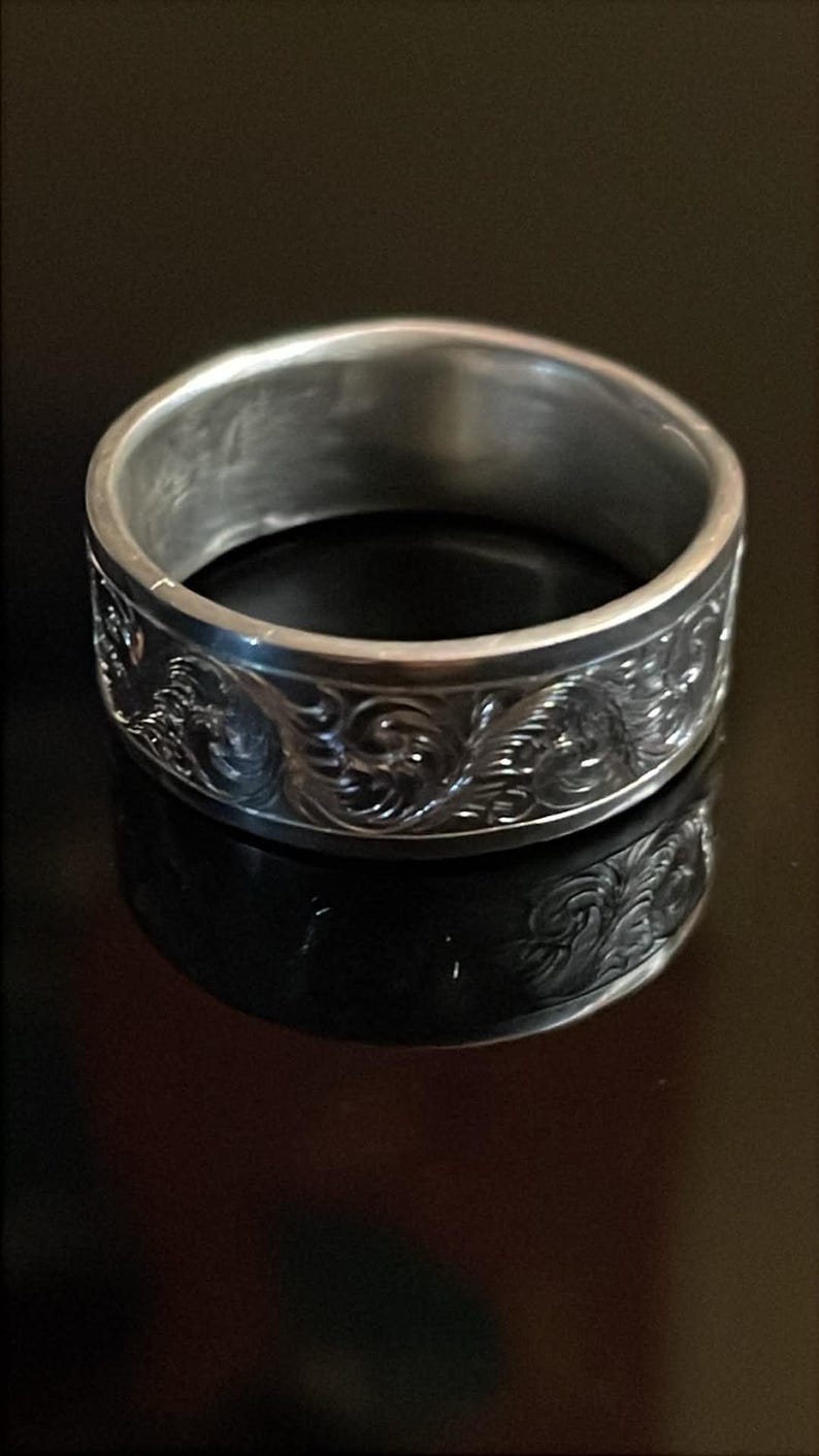 May include: A silver-toned metal ring featuring a continuous band with an intricate, embossed scrollwork pattern wrapping around the exterior surface. The ring has a smooth, polished interior and a reflective finish that catches the light.