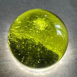 Puede incluir: Una canica de vidrio verde con un patrón de mármol en espiral. La canica es translúcida y tiene un acabado brillante.
