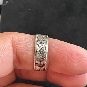 May include: A silver ring featuring an engraved floral design. The ring, displayed on a finger, showcases a detailed pattern around the band. The surface of the ring reflects light, emphasizing the intricate carvings.