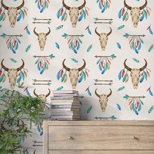 Könnte beinhalten: Tapete mit einem sich wiederholenden Muster aus Aquarell-Stierschädeln, Pfeilen und bunten Federn in Blau, Rot und Grün auf cremefarbenem Hintergrund. Eine Holzkommode und eine Pflanze befinden sich im Vordergrund.
