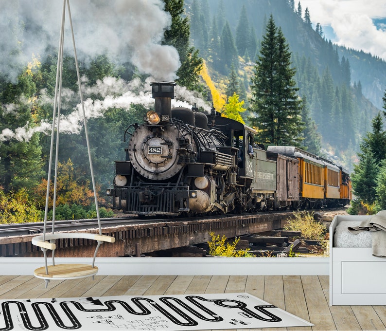May include: A wall mural depicting a black steam locomotive numbered 482 travelling across a wooden trestle bridge through a dense forest with tall pine trees. A white swing hangs in the foreground, and a white rug with a black road pattern lies below.