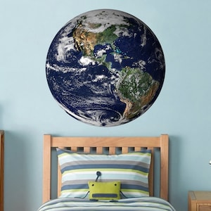 Peut inclure: Un autocollant mural représentant un globe terrestre bleu et vert avec une image détaillée de la Terre. L'autocollant est sur un mur bleu clair au-dessus d'un lit en bois avec un oreiller rayé et un jouet en peluche de grenouille verte.