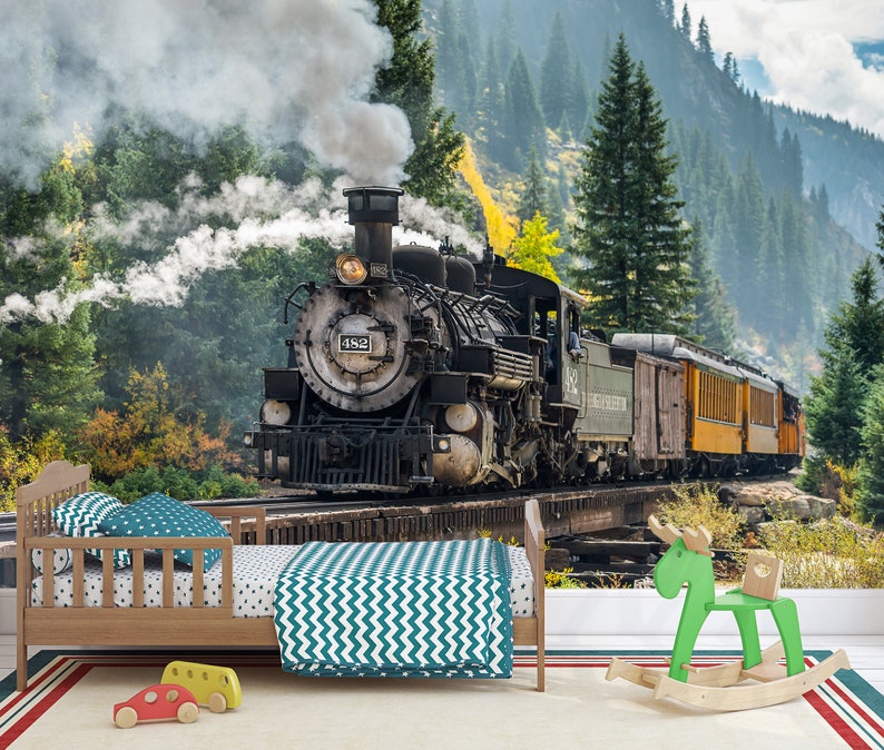 May include: A wooden toddler bed with a teal chevron blanket and polka dot pillows, positioned in front of a wall mural depicting a black steam locomotive numbered 482 on a bridge. A green wooden rocking moose and two small toy cars are on the floor.