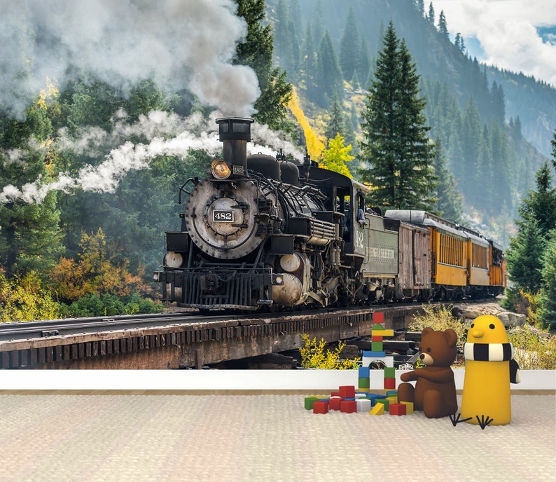 May include: A wall mural featuring a black steam locomotive numbered 482 travelling on a wooden trestle bridge through a forest with mountains in the background. In the foreground, a brown teddy bear, a yellow bird figure, and colourful building blocks.