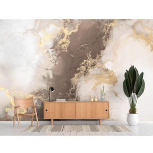 Puede incluir: Un interior moderno con una pared con efecto mármol en tonos blancos, marrones y dorados. Una silla de madera, un mueble bajo, una alfombra y una planta en maceta con grandes hojas verdes completan la decoración. Una lámpara de escritorio negra está sobre el mueble.