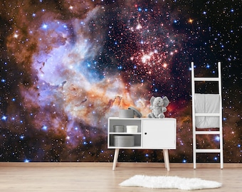 May include: A white bookshelf with a gray blanket on the top shelf. A gray elephant toy sits on a white shelf in front of a dark blue, orange, and purple nebula wallpaper with stars.