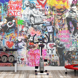 Op de afbeelding: Een stationaire hometrainer staat voor een muur bedekt met levendige graffitikunst met diverse figuren, stripfiguren en teksten zoals "Always Dream Big" en "I Love NY". Een halterrek en een kleine rode trampoline zijn ook aanwezig.