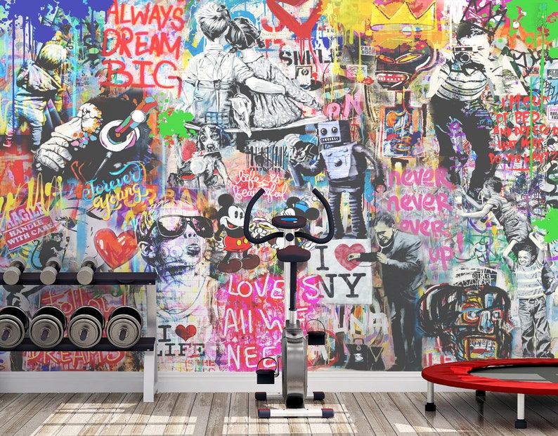 Op de afbeelding: Een stationaire hometrainer staat voor een muur bedekt met levendige graffitikunst met diverse figuren, stripfiguren en teksten zoals "Always Dream Big" en "I Love NY". Een halterrek en een kleine rode trampoline zijn ook aanwezig.